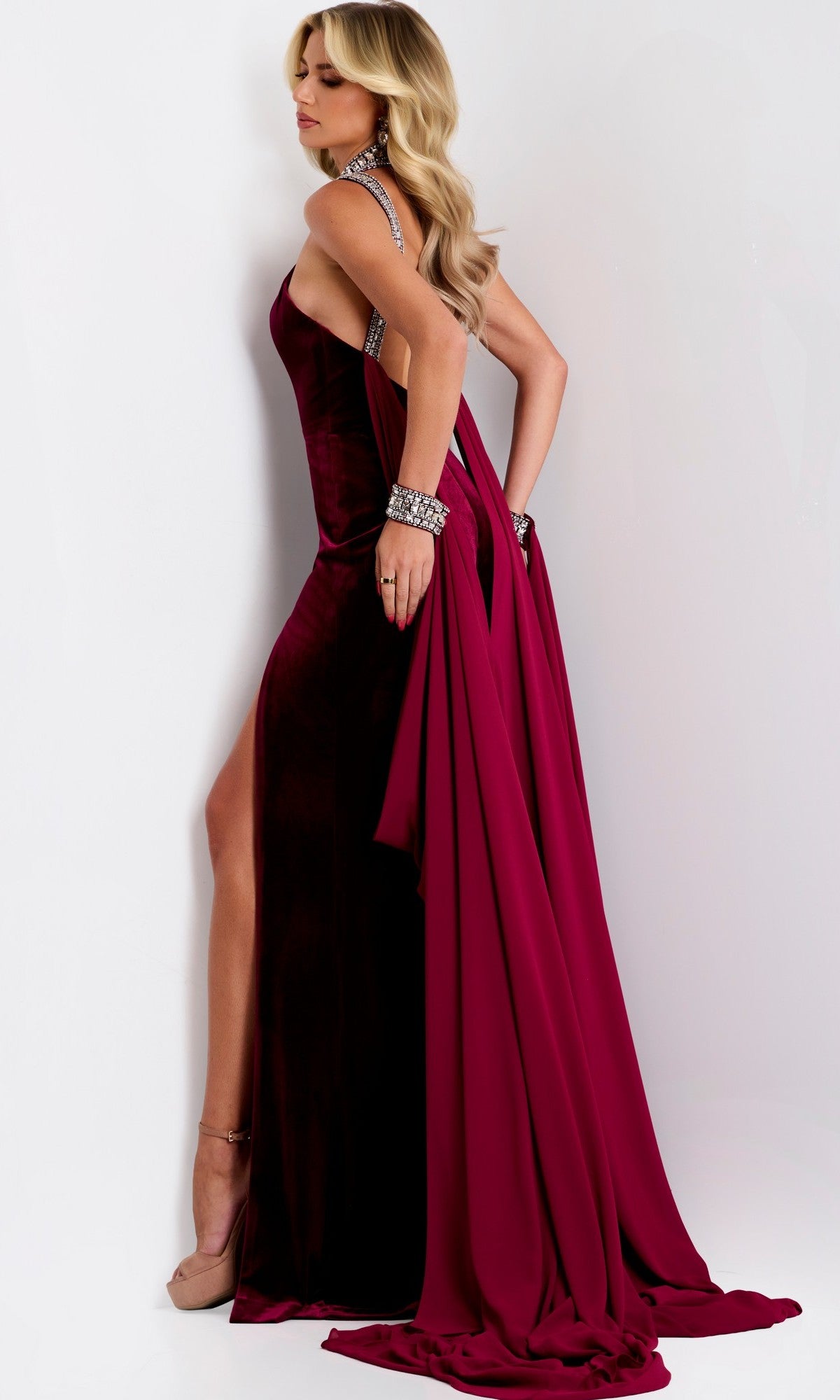 A woman wears the Jovani 43699 Long Prom Dress—a floor-length burgundy velvet halter gown with a high-leg slit and draped fabric—posing against a white background in heels and silver jewelry.